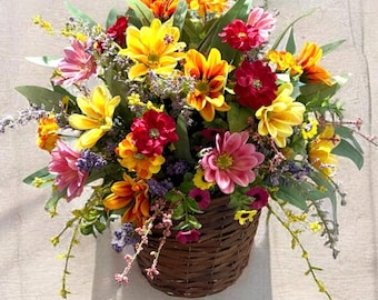 May include: A vibrant floral arrangement in a woven basket. The bouquet features yellow, red, pink, and orange flowers with green foliage. The basket is brown and hangs against a light-colored wall, creating a cheerful display.