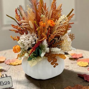 May include: A white pumpkin-shaped container filled with an arrangement of dried and faux botanical elements in shades of orange, cream, and brown, including small orange flowers, feathery grasses, and textured foliage stems.