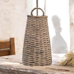 May include: A tall, woven basket with a handle. The basket is made of light brown wicker and has a tapered shape. The handle is a twisted rope design. The basket is sitting on a wooden table.