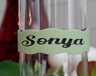 May include: A clear glass vase with a green label that says "Sonya" in black cursive lettering.