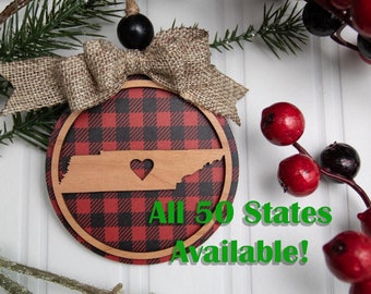 Puede incluir: Un adorno navideño de cuadros rojos y negros con un recorte de madera del estado de Tennessee, con un corazón en el centro. El adorno está decorado con un lazo de arpillera y una cuenta negra. El texto en el adorno dice "¡Todos los 50 estados disponibles!"
