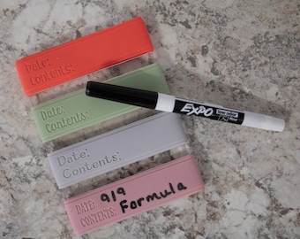 May include: Four silicone food storage containers with labels for date and contents. The containers are red, green, gray, and pink. A white dry-erase marker is lying on top of the containers.