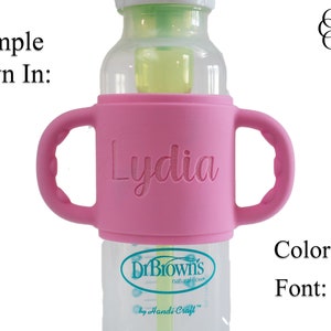 May include: Pink silicone bottle holder with two handles, personalised with the name "Lydia" in a Chloe font. The holder fits a Dr. Brown's bottle.