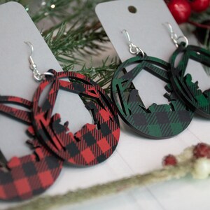 May include: Two pairs of Christmas-themed earrings. One pair features a red and black plaid design with a cutout of the nativity scene. The other pair features a green and black plaid design with a cutout of the nativity scene.