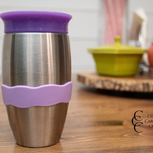 Puede incluir: Una taza de viaje de acero inoxidable con una tapa morada y una banda de silicona morada alrededor del medio. La taza est&aacute; sobre una mesa de madera.
