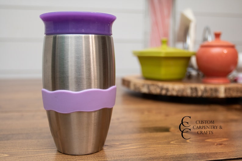 Puede incluir: Una taza de viaje de acero inoxidable con una tapa morada y una banda de silicona morada alrededor del medio. La taza est&aacute; sobre una mesa de madera.