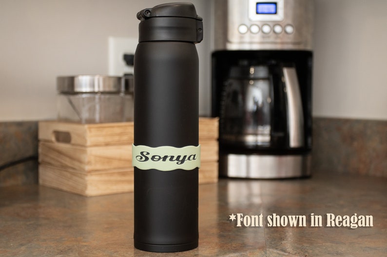May include: Black stainless steel water bottle with a green personalized label that says "Sonya". The font is shown in Reagan.