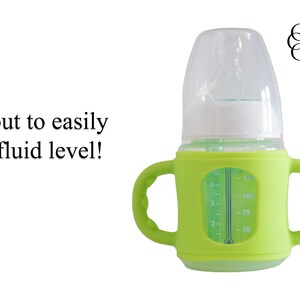 May include: A green silicone baby bottle with a clear glass insert and a white lid. The bottle has a handle on each side and a measurement scale on the side of the glass insert. The text "Cut out to easily see fluid level!" is visible in the image.