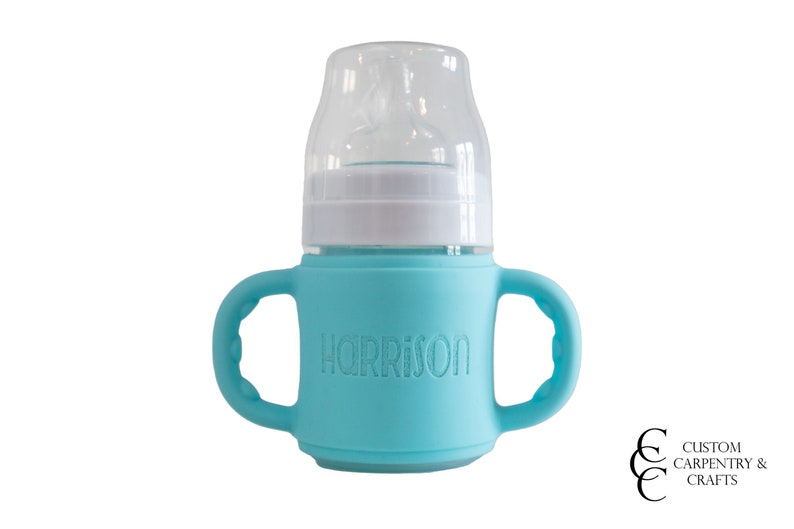 May include: A turquoise blue silicone baby bottle with two handles and a clear plastic lid. The bottle has the name "HARRISON" embossed on the front.