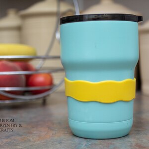 Puede incluir: Una taza de viaje azul claro con una banda de silicona amarilla alrededor del medio. La taza tiene una tapa negra y una pajita.