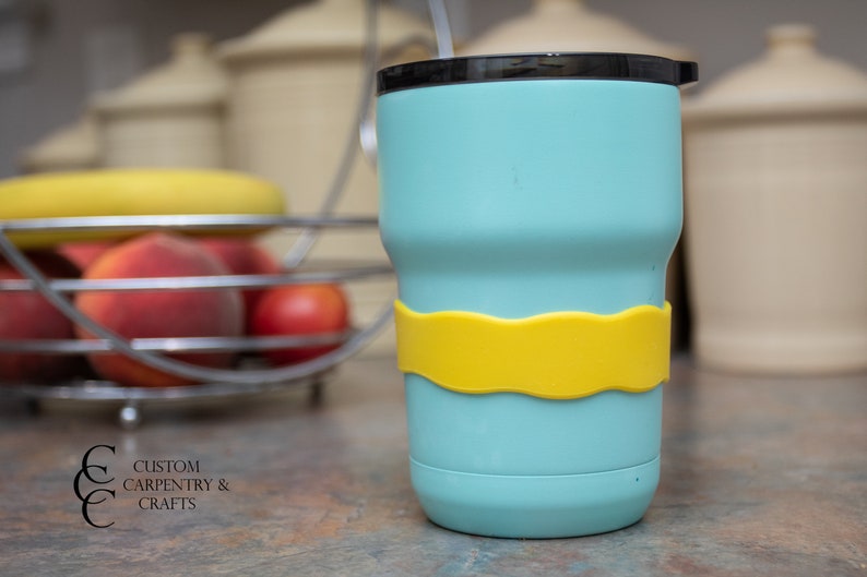 Puede incluir: Una taza de viaje azul claro con una banda de silicona amarilla alrededor del medio. La taza tiene una tapa negra y una pajita.