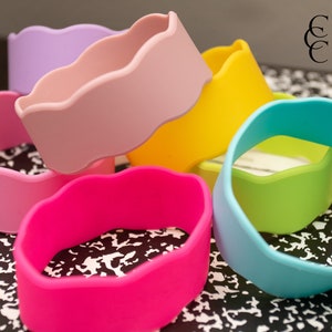 Puede incluir: Seis pulseras de silicona de colores con un borde festoneado. Las pulseras est&aacute;n apiladas una encima de la otra y est&aacute;n dispuestas en un patr&oacute;n de arco&iacute;ris. Las pulseras est&aacute;n sobre una superficie con un patr&oacute;n blanco y negro.