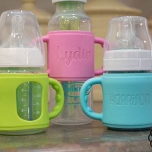 May include: Three baby bottles with personalised silicone sleeves. The bottles are clear glass with a white nipple and a green, pink, and blue silicone sleeve. The sleeves have the names "Lydia", "Harrison", and a measurement scale.