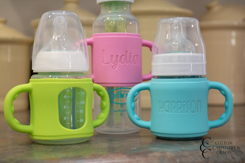 May include: Three baby bottles with personalised silicone sleeves. The bottles are clear glass with a white nipple and a green, pink, and blue silicone sleeve. The sleeves have the names "Lydia", "Harrison", and a measurement scale.