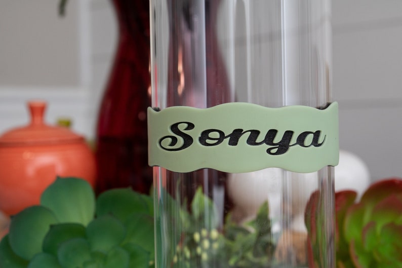May include: A clear glass vase with a green label that says "Sonya" in black cursive lettering.