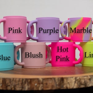May include: A set of eight silicone cups with handles in various colours, including pink, purple, marble, blue, blush, hot pink, and lime. The cups are stacked on a wooden surface.