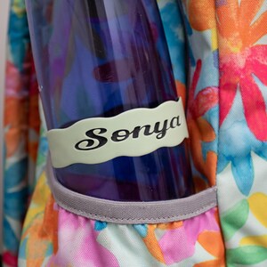 May include: A blue water bottle with a light green personalized name tag that says "Sonya" in black lettering. The bottle is in a floral patterned backpack.