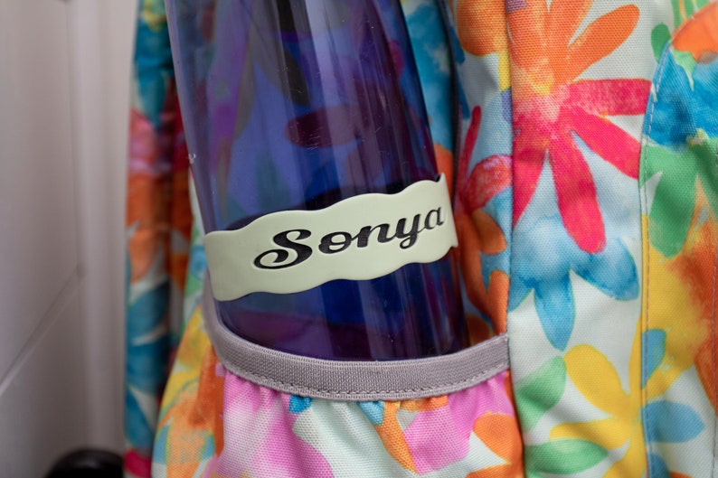 May include: A blue water bottle with a light green personalized name tag that says "Sonya" in black lettering. The bottle is in a floral patterned backpack.