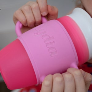 May include: A pink and white silicone sippy cup with a handle and the word "yodia" embossed on the side. The cup is being held by a child.
