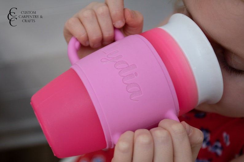 May include: A pink and white silicone sippy cup with a handle and the word "yodia" embossed on the side. The cup is being held by a child.