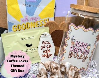 May include: A gift box filled with coffee-themed items, including a glass jar with a straw, a white keychain with the text "let's go get iced coffee", a gold eye mask, and a brown hair scrunchie. The box is decorated with the text "Mystery Coffee Lover Themed Gift Box".