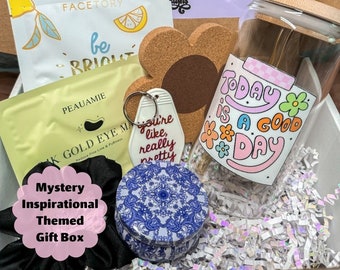 May include: A gift box filled with various items, including a face mask, a keychain, a candle, a hair scrunchie, a cork coaster, and a glass jar with a straw. The box is decorated with a pink and white label that says "Mystery Inspirational Themed Gift Box".