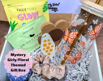 May include: A white gift box filled with various items, including a face mask, a glass tumbler with a butterfly design, a key chain, a hair scrunchie, and a cork heart. The box is labeled "Mystery Girly/Floral Themed Gift Box".