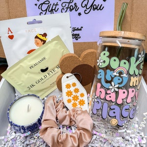 May include: A gift box filled with various items, including a candle, a face mask, a keychain, a hair scrunchie, a glass with a "Books are my happy place" design, and a straw. The box is lined with white tissue paper.