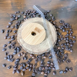 May include: A wooden lid with a hole, a clear glass straw, and a wooden coaster are surrounded by roasted coffee beans on a wooden surface. The lid and coaster are made of light-colored wood.
