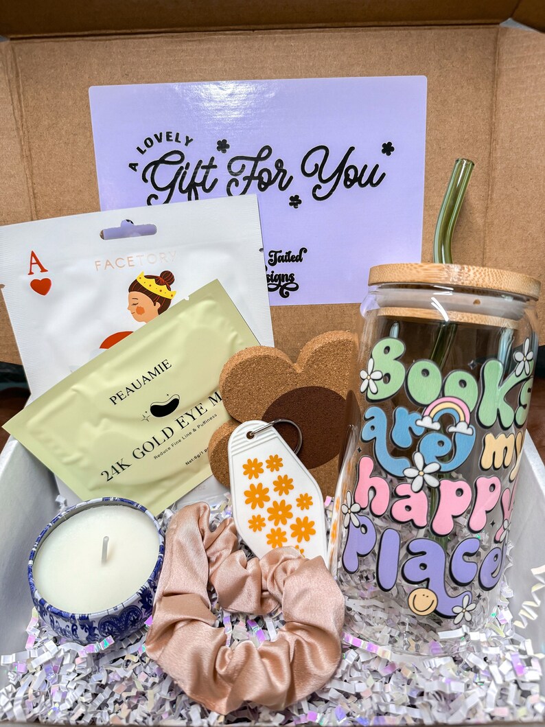 May include: A gift box filled with various items, including a candle, a face mask, a keychain, a hair scrunchie, a glass with a "Books are my happy place" design, and a straw. The box is lined with white tissue paper.
