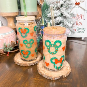 May include: Two clear glass cups with lids and straws, decorated with green and red Christmas wreaths and Mickey Mouse ears. The cups are filled with a light brown liquid and are sitting on wooden coasters.