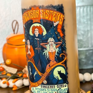 May include: A glass tumbler with a bamboo lid, filled with a light brown beverage. The tumbler features a colorful graphic of the Sanderson Sisters from the movie, with text that reads "The Sanderson Sisters Live From The Dead". Additional text includes "October 31-1993" and "Salem, Mass".