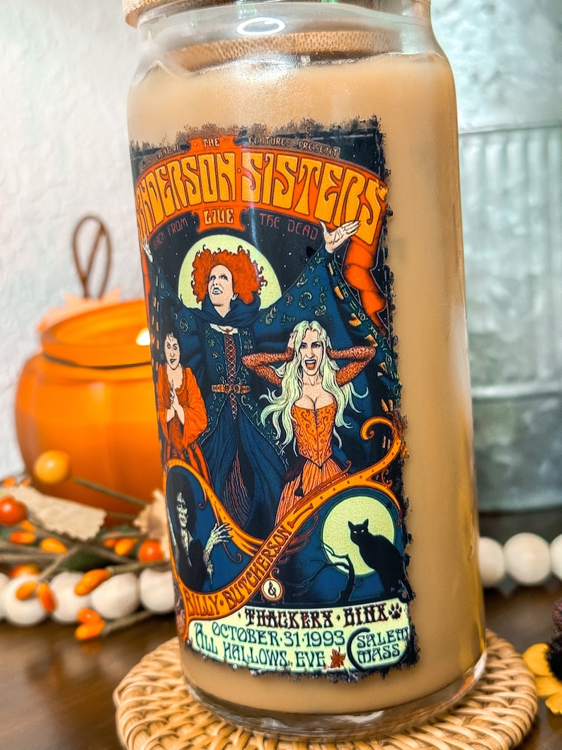 May include: A glass tumbler with a bamboo lid, filled with a light brown beverage. The tumbler features a colorful graphic of the Sanderson Sisters from the movie, with text that reads "The Sanderson Sisters Live From The Dead". Additional text includes "October 31-1993" and "Salem, Mass".