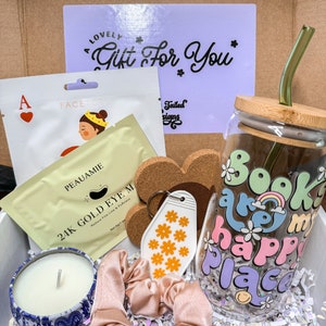 May include: A gift box filled with various items, including a glass tumbler with the text "Books are my happy place" in rainbow colors, a candle in a blue and white tin, a pink hair scrunchie, a key chain with a white tag decorated with orange flowers, a face mask, and a cork coaster with the text "Tailed Designs".