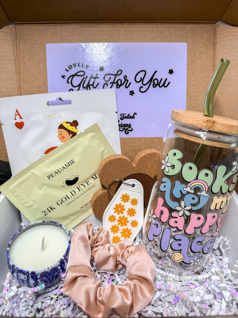 May include: A gift box filled with various items, including a glass tumbler with the text "Books are my happy place" in rainbow colors, a candle in a blue and white tin, a pink hair scrunchie, a key chain with a white tag decorated with orange flowers, a face mask, and a cork coaster with the text "Tailed Designs".