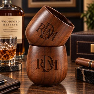 May include: Two dark brown wooden cups with a smooth, rounded shape, each featuring a laser-engraved monogram of the letters RDM centered on the front. The wood grain is visible throughout the surface of both cups, which are stacked vertically.