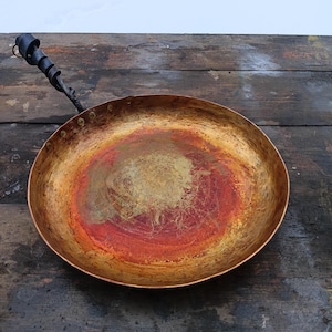 May include: A copper-colored, round frying pan with a dark, twisted metal handle. The pan's interior has a reddish-orange and gold patina, suggesting use. The pan is set against a rustic, weathered wooden surface.