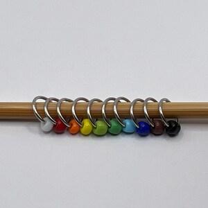 May include: A set of ten colorful stitch markers on a bamboo knitting needle. The markers are made of metal rings with glass beads in various colors, including white, red, orange, yellow, green, blue, dark blue, brown, and black.