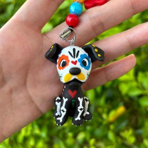 Puede incluir: Una pequeña figurita de perro blanco y negro con una cara de calavera de azúcar del Día de Muertos. La figurita tiene flores coloridas y un corazón en el pecho. La figurita está colgada de un collar de cuentas de colores.