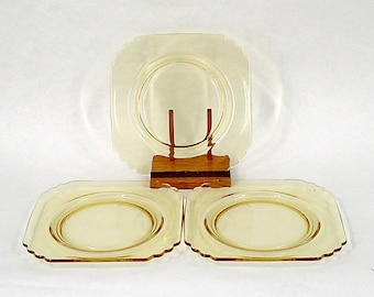 Puede incluir: Un juego de tres platos de vidrio amarillo vintage. Los platos tienen forma cuadrada con bordes festoneados y un diseño circular en el centro. Un plato se exhibe en un soporte de madera. Los platos son de color amarillo claro.