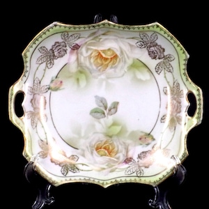 May include: A white porcelain serving dish with gold trim and a floral design featuring white roses with yellow centers. The dish has two handles and a scalloped edge.