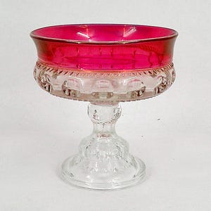 May include: A clear glass footed bowl with a vibrant red rim. The bowl's upper section is a deep red, transitioning to a clear, patterned lower section. The stem and base are also clear glass, showcasing intricate detailing. A decorative piece.