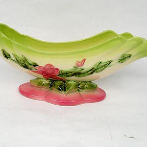 Può includere: Un vaso in ceramica a forma di barca, con esterno verde chiaro e interno color crema. Il vaso è decorato con fiori rosa e foglie verdi e poggia su una base rosa a forma di foglia. Un pezzo decorativo.
