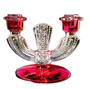 May include: A clear glass double candlestick holder with ruby red accents. The base and candle cups are ruby red, while the arms and central column are clear, with a decorative, geometric design. The candlestick holder is designed to hold two candles.