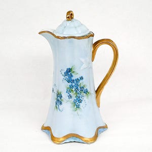 Könnte beinhalten: Eine hellblaue Kaffeekanne aus Porzellan mit einem handgemalten Blumenmuster aus kleinen blauen Blüten und grünen Blättern. Das Gefäß hat einen gewellten Boden und Rand mit Goldverzierungen sowie einen passenden Deckel mit goldenem Knauf.