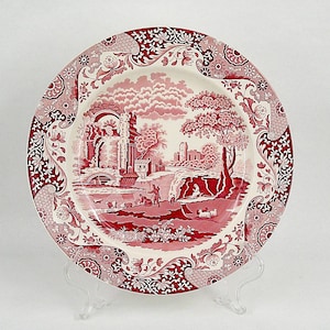 Op de afbeelding: Een ronde plaat met een rood en wit landschapsontwerp. De plaat heeft een gedetailleerde scène met gebouwen, bomen en figuren, omlijst door een decoratieve rand. De plaat wordt gepresenteerd op een transparante standaard.