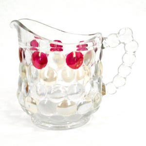 May include: A clear glass jug with a distinctive bubble pattern. The handle is formed from joined glass spheres. The body of the jug is adorned with alternating red and white circular accents, giving it a decorative and appealing look.