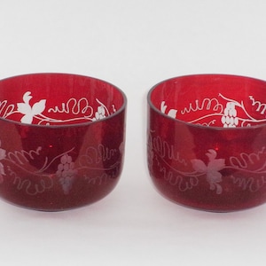 Może przedstawiać: Dwa rubinowo-czerwone szklane kubki z białym, wytrawionym wzorem winorośli. Szklanki są cylindryczne i mają gładkie, polerowane wykończenie. Wzór winorośli obejmuje liście i kiście winogron.