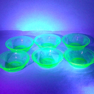 May include: Six small, clear glass bowls with a bright neon green glow. The bowls have a rounded shape with a slightly flared rim and a decorative pattern around the edge. The bowls are arranged on a purple surface.