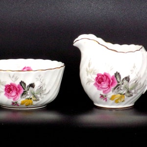 Może przedstawiać: Biały porcelanowy zestaw do śmietanki i cukru. Oba elementy zdobi różowy wzór kwiatowy z różami ze złotym wykończeniem. Cukiernica ma spiralny wzór, a śmietanka uchwyt.